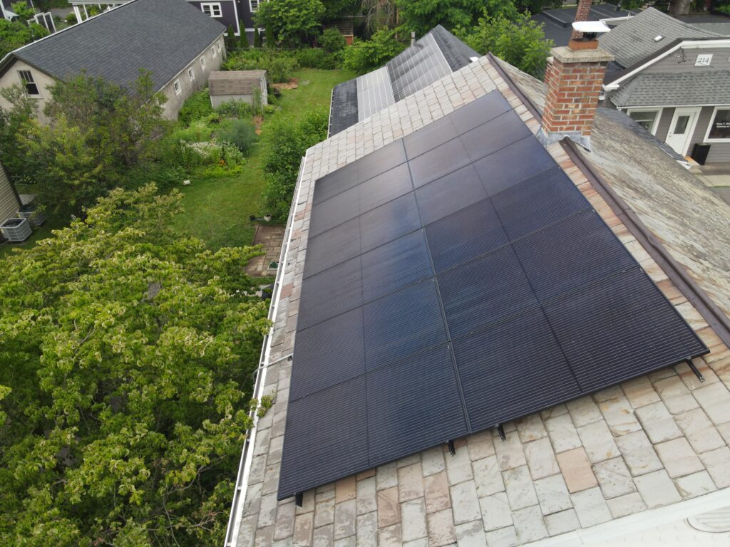 Aerial view of a black solar array mounted on the gray slate roof of a home in Northampton, Massachusetts, beside a brick chimney.