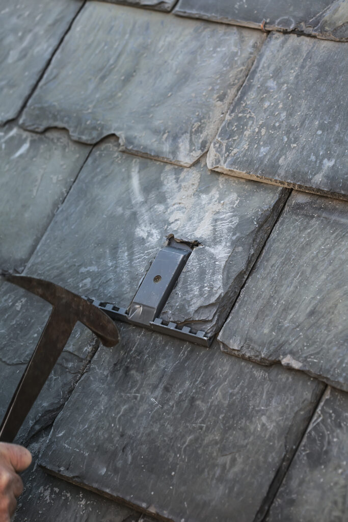Close-up of a SlatePlate metal mounting bracket installed flush between gray slate tiles, with a slate ripping tool alongside.
