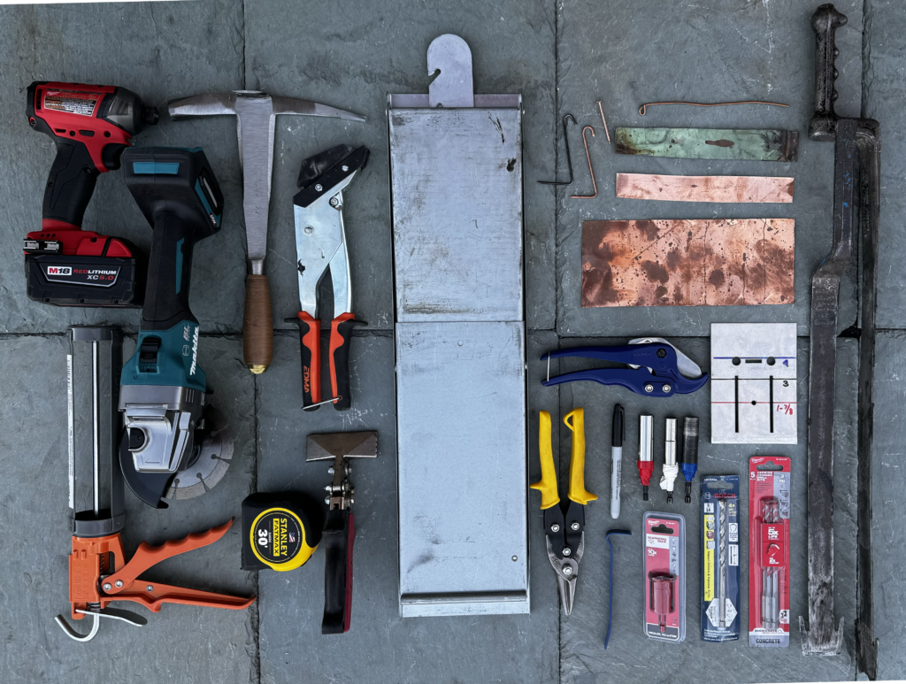 Overhead flat-lay of slate-roof solar installation tools and materials on slate, including a cordless impact driver, angle grinder with diamond blade, slate hammer, slate cutter, caulk gun, tin snips, tape measure, and copper and metal flashing.
