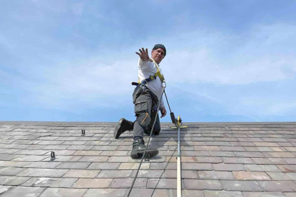 A roofer kneeling on a slate roof reaching toward the camera, wearing a safety harness, with SlatePlate mounting brackets installed on the slate and a bright blue sky behind.